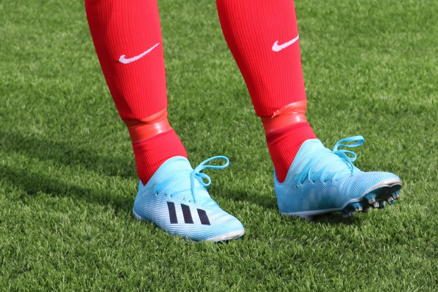 Beine in roten Stutzen und hellblauen Fußballschuhen mit Stollen auf kurzflorigem Kunstrasen