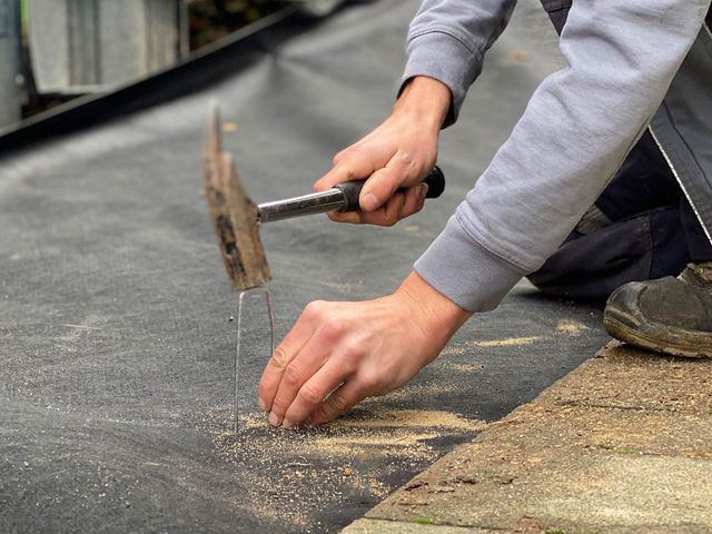 Ein Erdnagel wird mit dem Hammer durch das Unkrautvlies in den Untergrund geschlagen