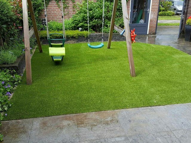 Kunstrasenfläche mit Schaukelgerüst und Nestschaukel, daneben nasses Pflaster und eine Hecke