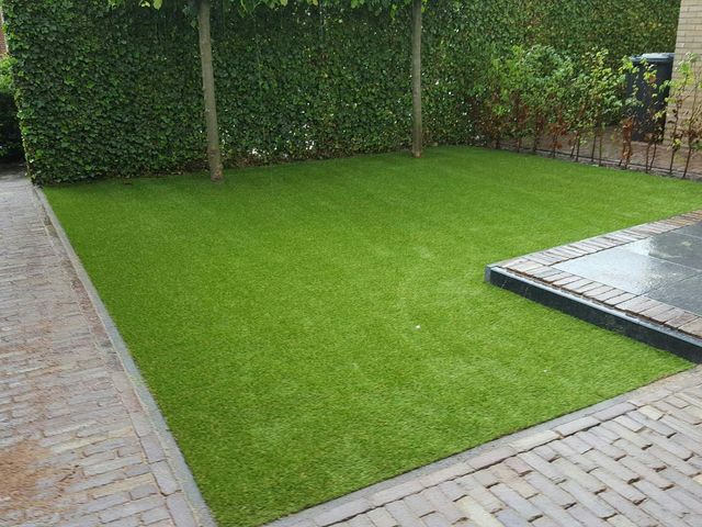 Rechteckige Kunstrasenfläche im Garten, eingefasst von einer hohen Hecke, mit Klinkerpflaster und Holzpfosten
