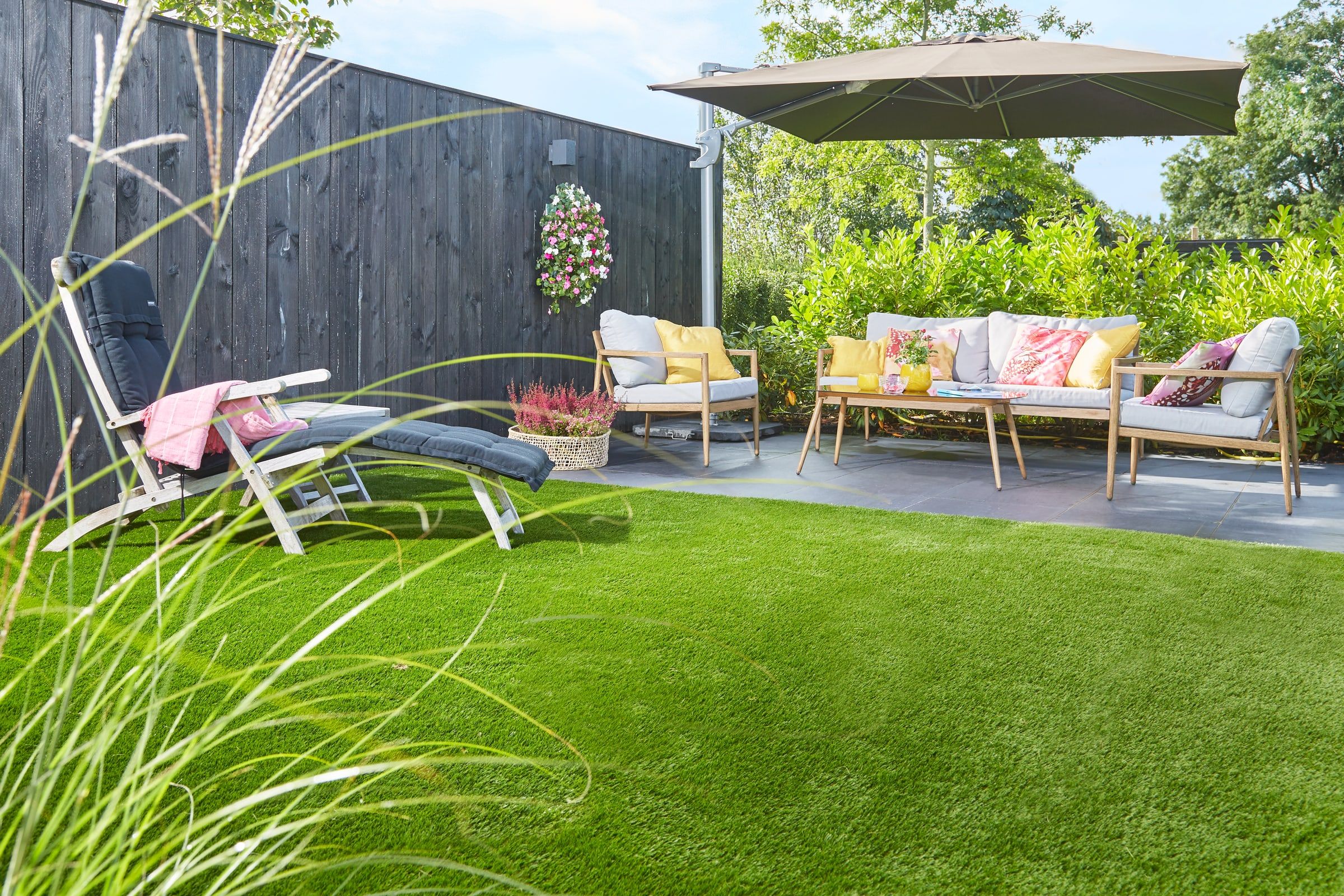 Garten mit Kunstrasenfläche, Loungegarnitur unter einem Sonnenschirm und Ziergras im Vordergrund