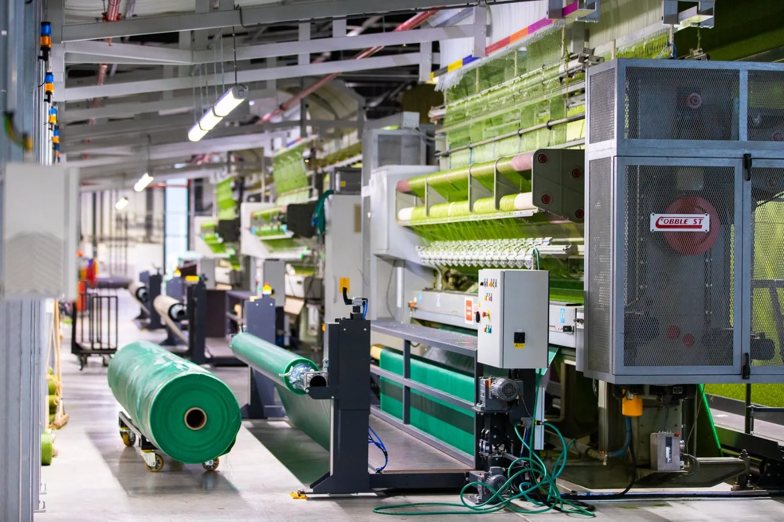Produktionshalle mit Tuftmaschinen, aufgespanntem grünem Garn und fertigen Rollen