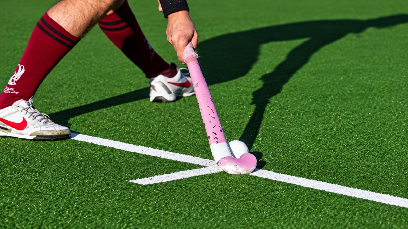 Hockeyschläger führt einen weißen Ball an einer Linienkreuzung auf kurzflorigem Kunstrasen, daneben die Füße des Spielers