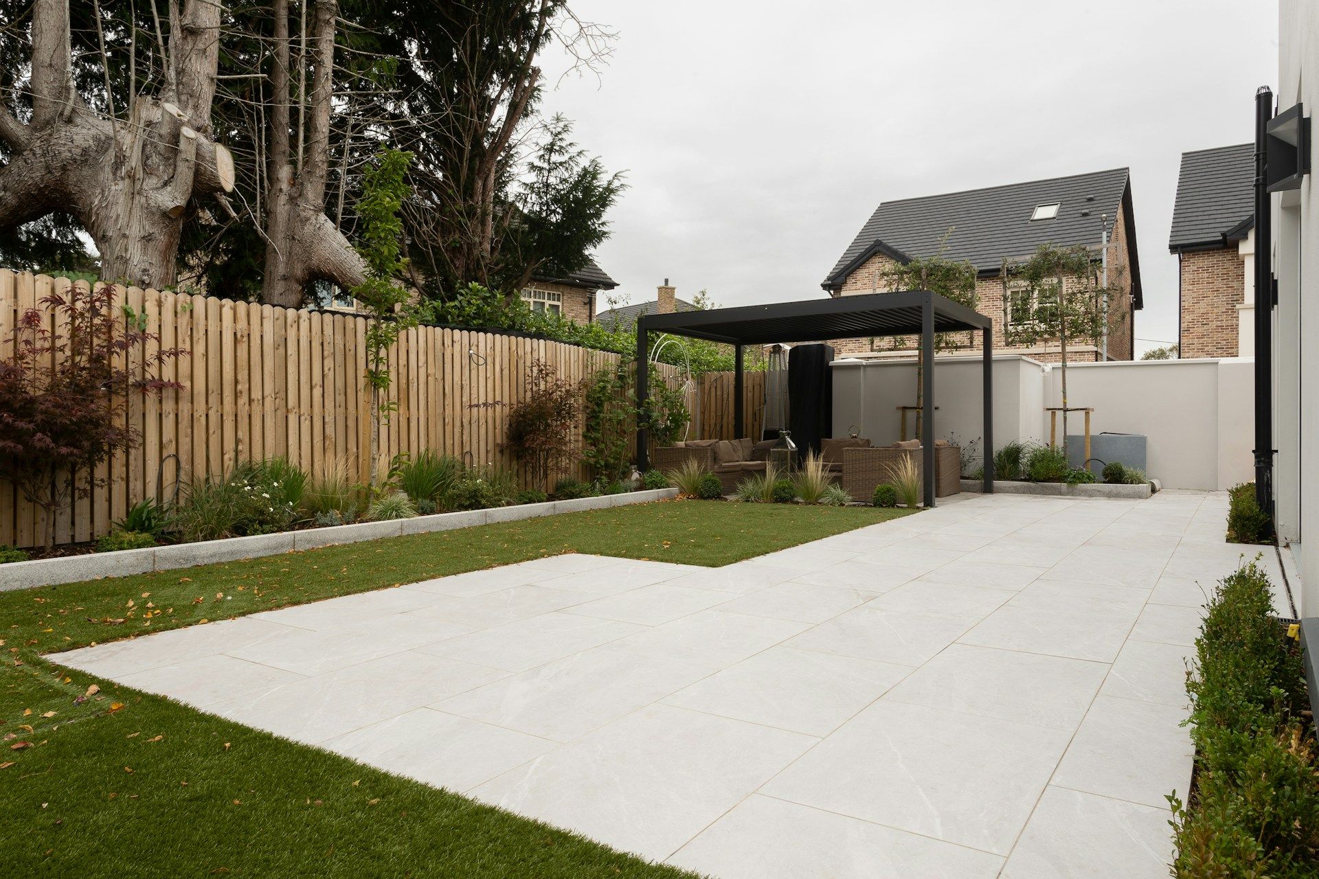 Garten mit großformatigen hellen Terrassenplatten, zwei Rasenflächen, Holzzaun und einer Pergola mit Loungemöbeln im Hintergrund