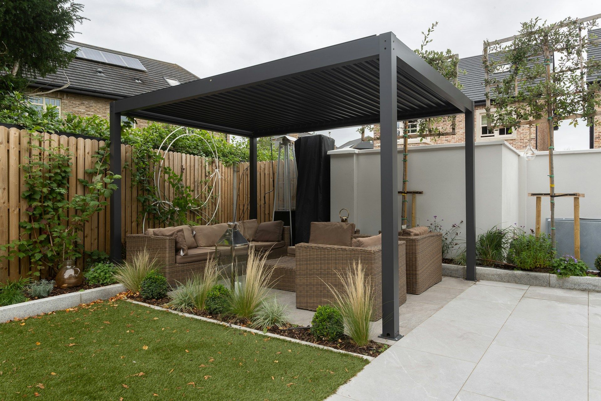 Pergola mit Rattan-Loungemöbeln auf einer Plattenterrasse, davor Ziergräser und eine Rasenfläche